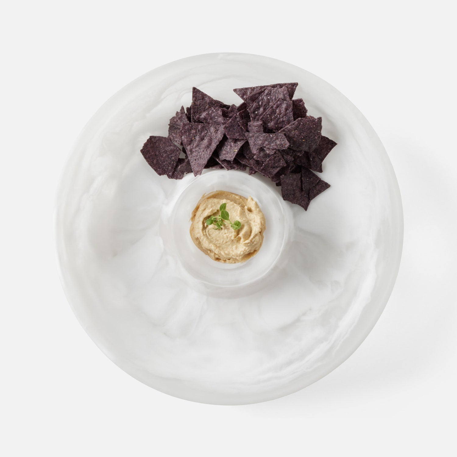 A white ceramic party platter with a central dip cup filled with hummus and garnished with a green herb, surrounded by dark purple blue corn tortilla chips on a white background.