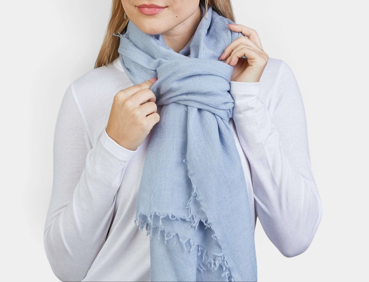 The Copper Shed arctic blue 100% Mongolian cashmere scarf with frayed edges worn by a woman in a white long-sleeve top.