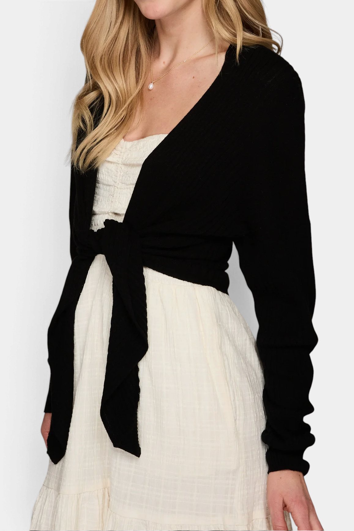 The Copper Shed black ribbed cashmere cardigan with a tie-front design worn over a cream dress