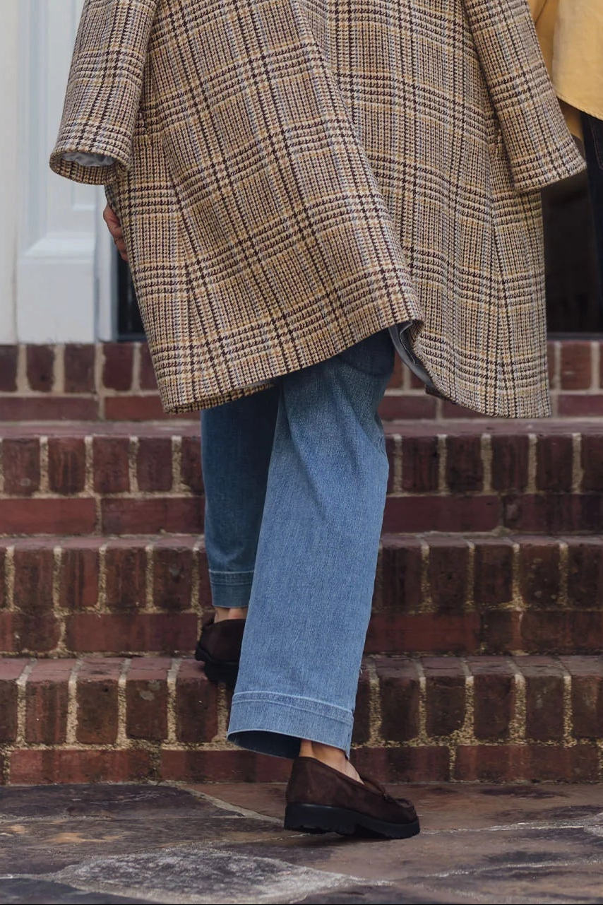 The Copper Shed brown suede lug sole loafers worn with blue jeans and a plaid coat while walking up brick stairs.
