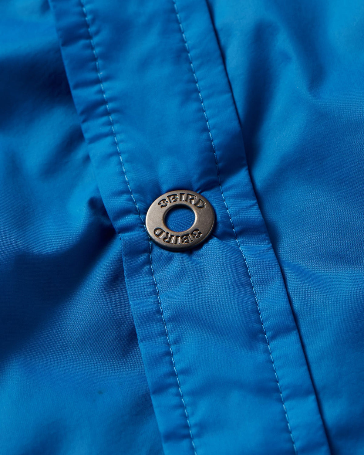 Close-up of a bright blue The Copper Shed Frigate Windshirt featuring a metallic button with engraved branding.