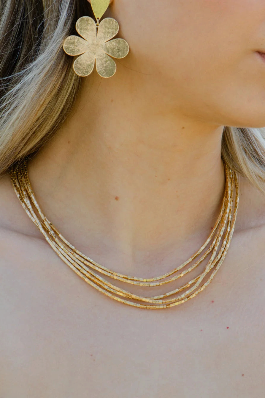 The Copper Shed Amunet 18k gold-plated multi-strand beaded necklace worn by a woman with gold flower earrings.