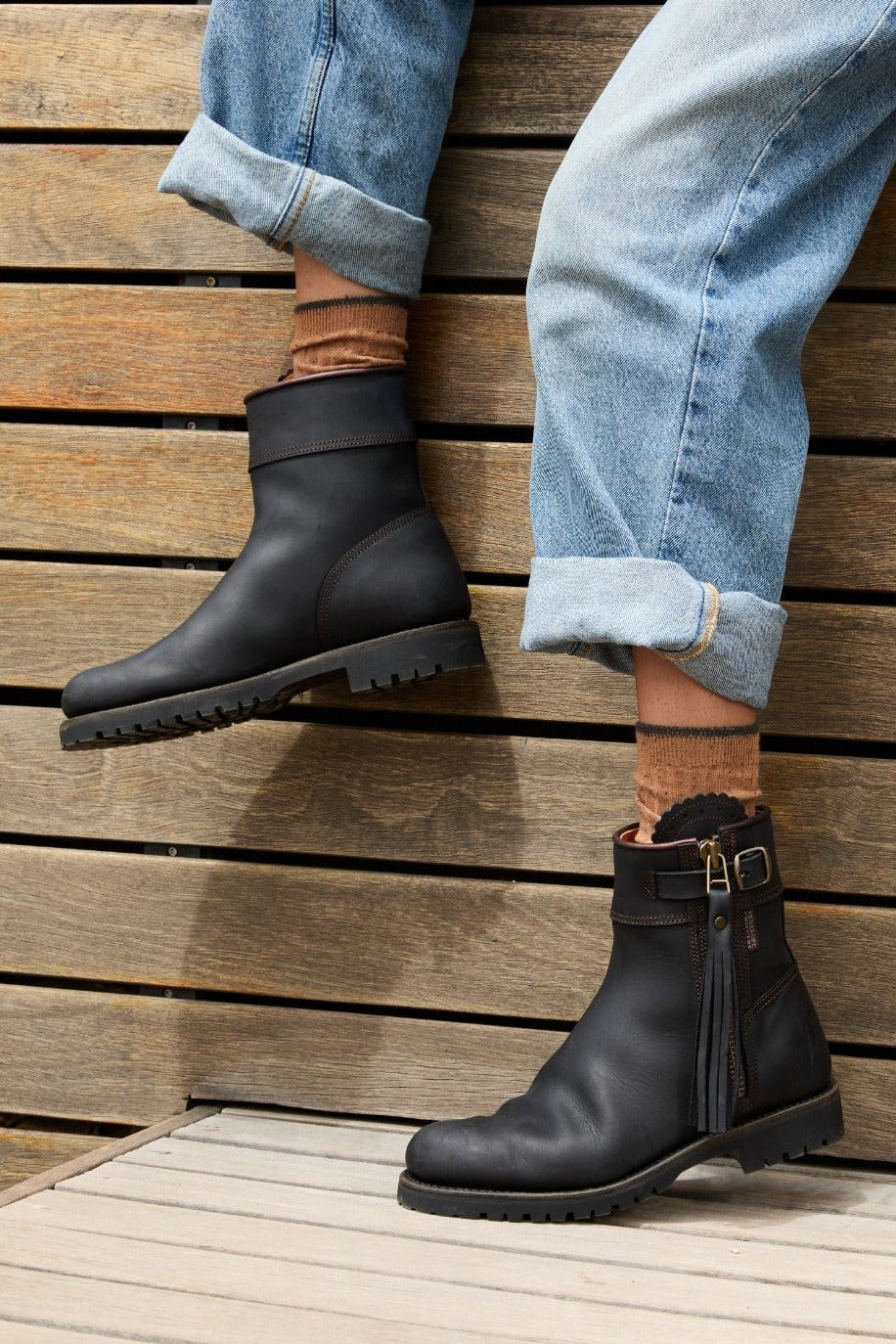 The Copper Shed Inclement cropped black waterproof suede boots with tassel detail and side buckle on a wooden deck.