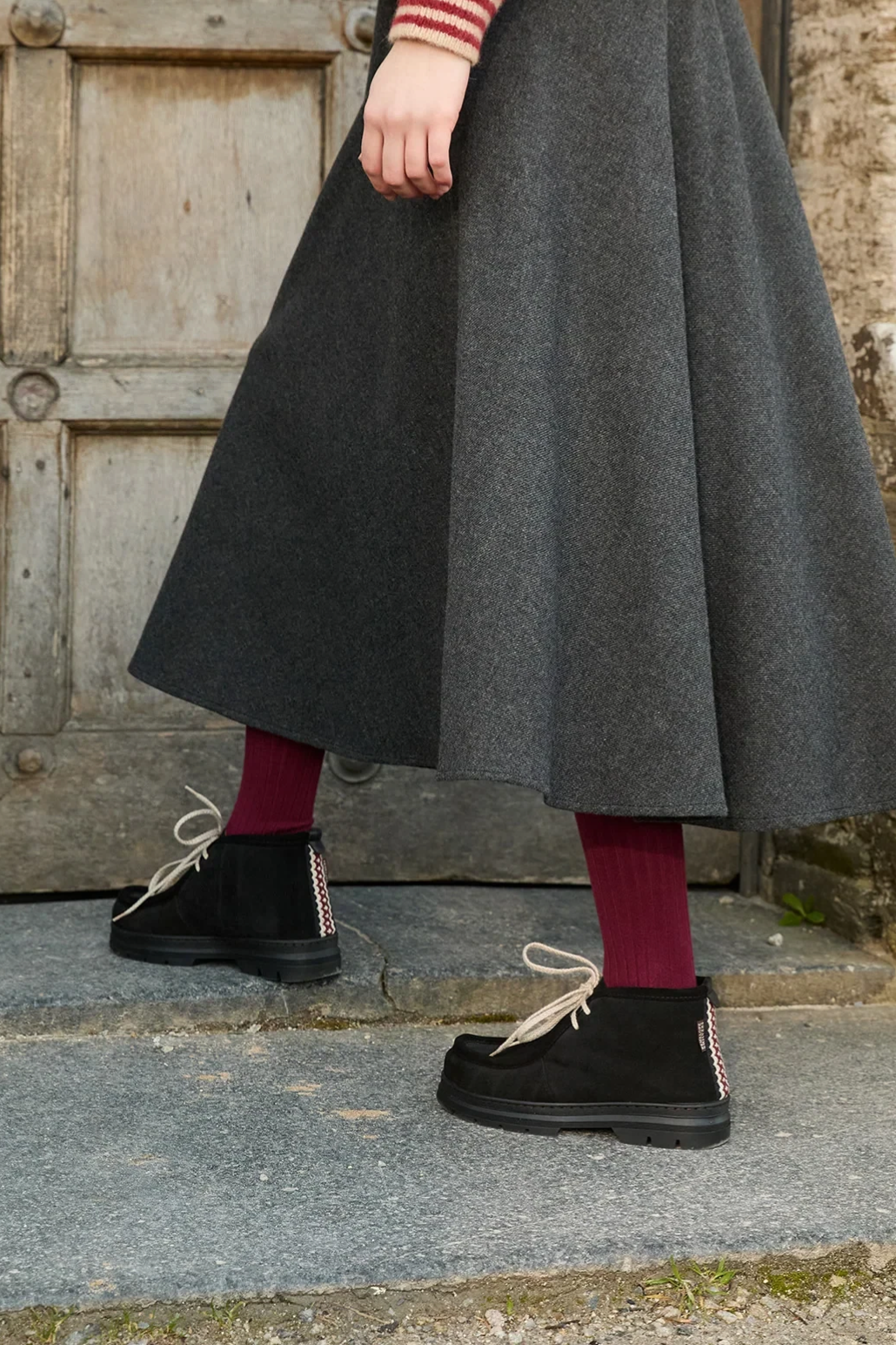 The Copper Shed black suede shearling-lined boots with ecru laces and folk trim detail worn with a grey midi skirt.
