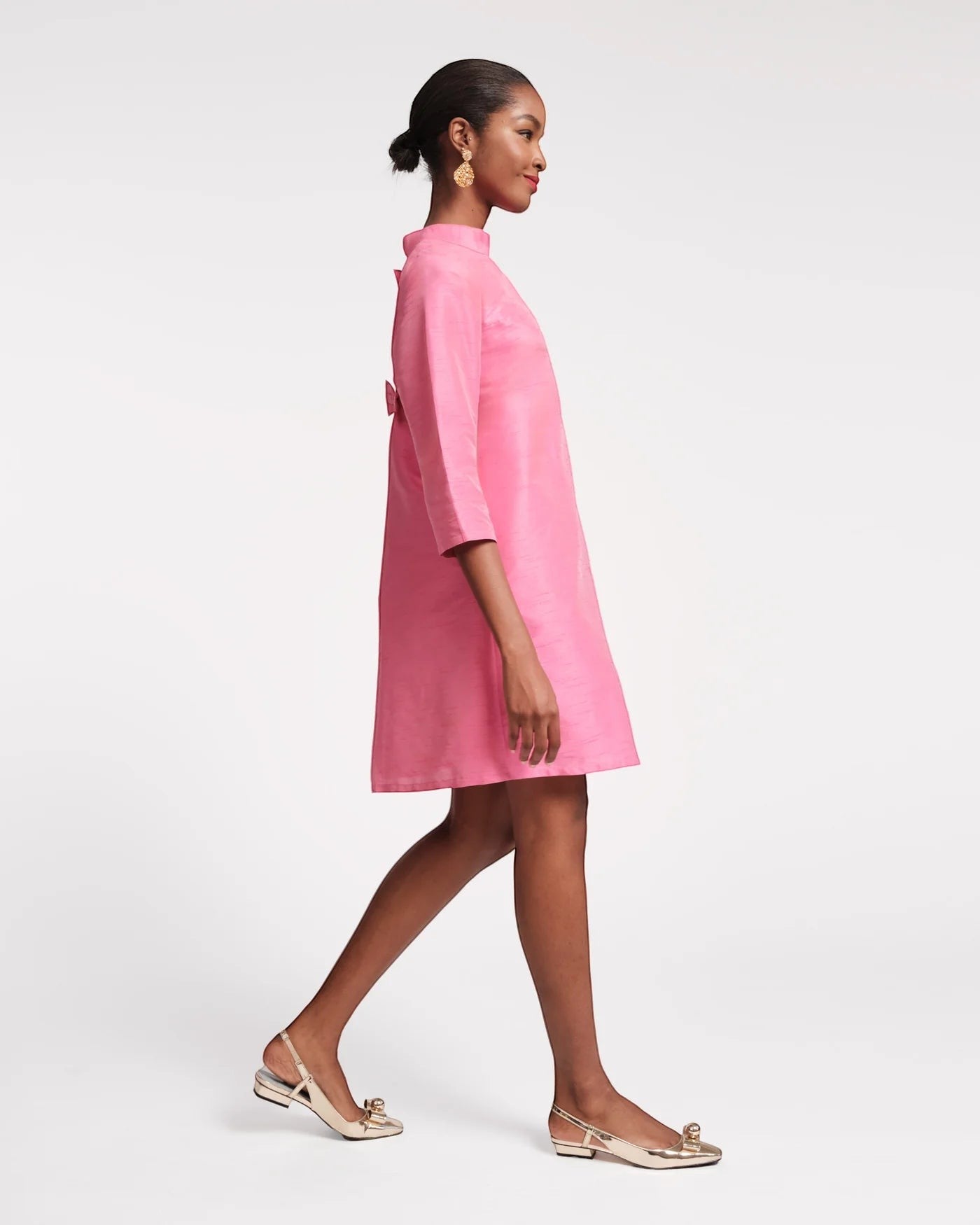 The Copper Shed pink mini dress with mock neckline, 3/4 sleeves, and bow detail on back. Model wears gold flats.