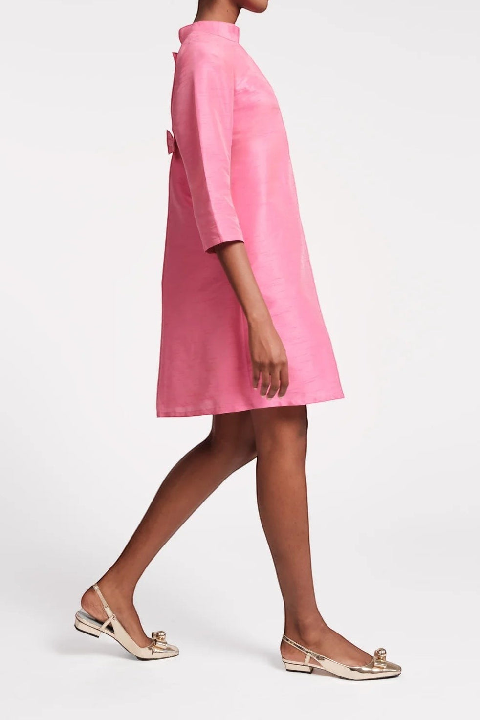The Copper Shed pink mini dress with mock neckline, 3/4 sleeves, and bow detail on back. Model wears gold flats.