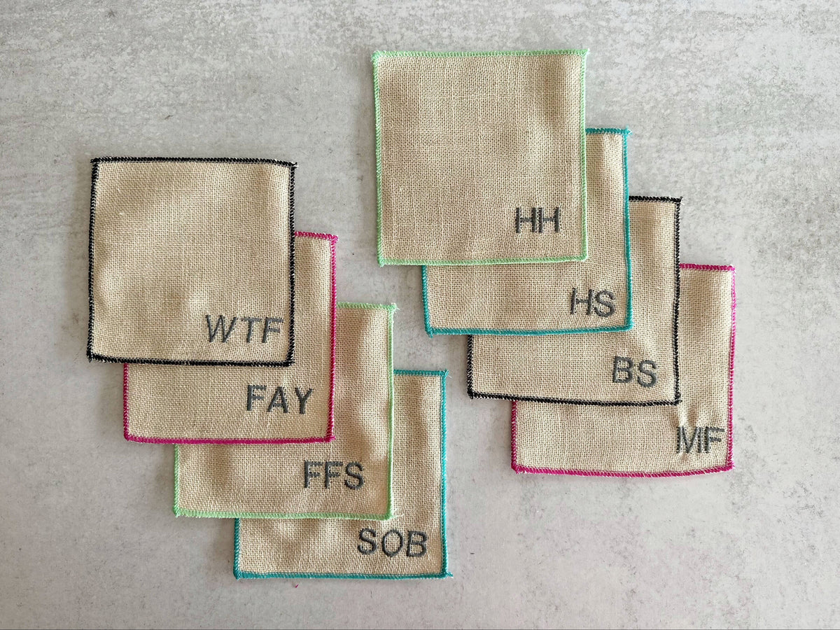 The Copper Shed set of 8 beige linen poly blend square cocktail coasters with colorful stitched edges and swear words.