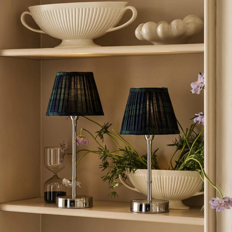 The Copper Shed pleated dark green tartan lampshades on silver rechargeable lamp bases displayed on a shelf.