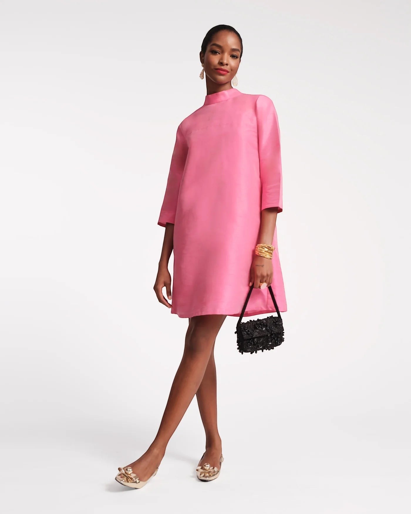 The Copper Shed Diplomat Mini Dress in bright pink silk with a mock neckline and 3/4 sleeves. Model wears gold earrings and bracelets.