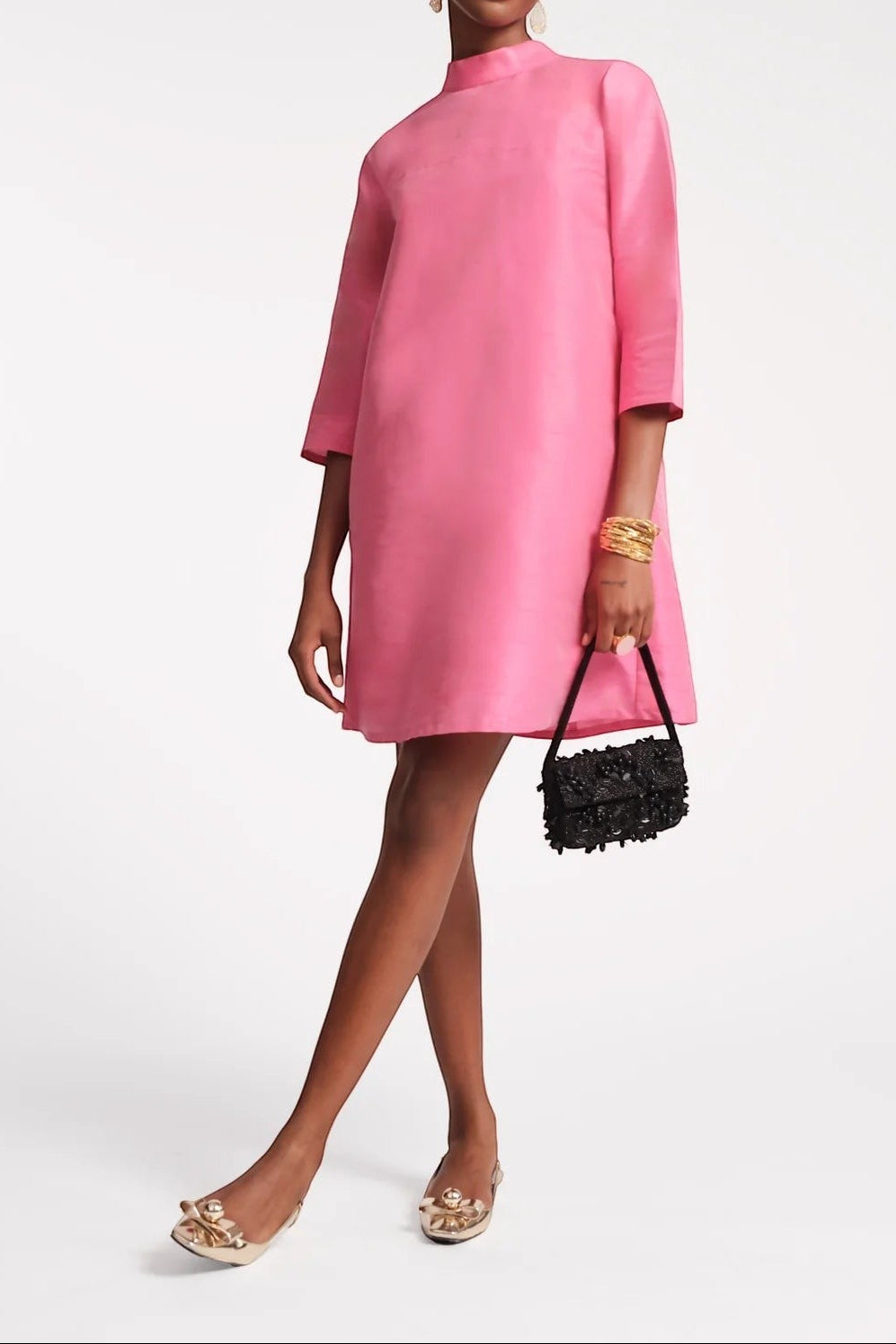 The Copper Shed Diplomat Mini Dress in bright pink silk with a mock neckline and 3/4 sleeves. Model wears gold earrings and bracelets.