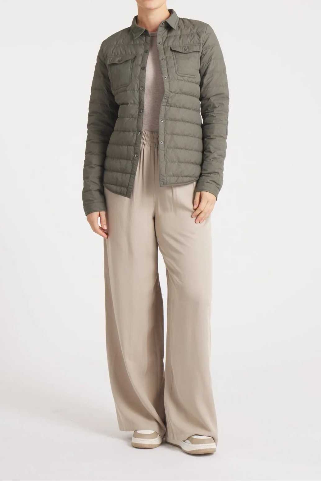 A woman modeling a dark olive green quilted puffer jacket over a beige top and wide-leg beige trousers, standing against a light gray studio background.