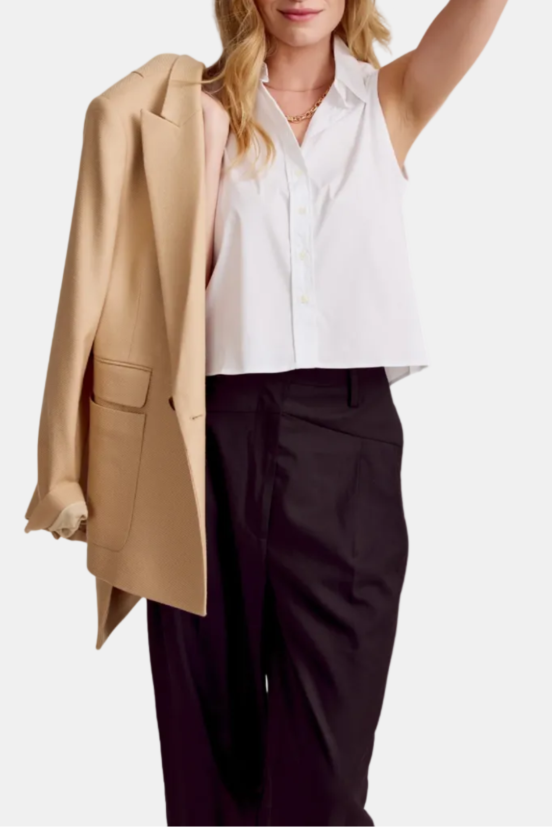 A woman wearing a beige sleeveless shirt with black pants. She has one hand raised to her head.