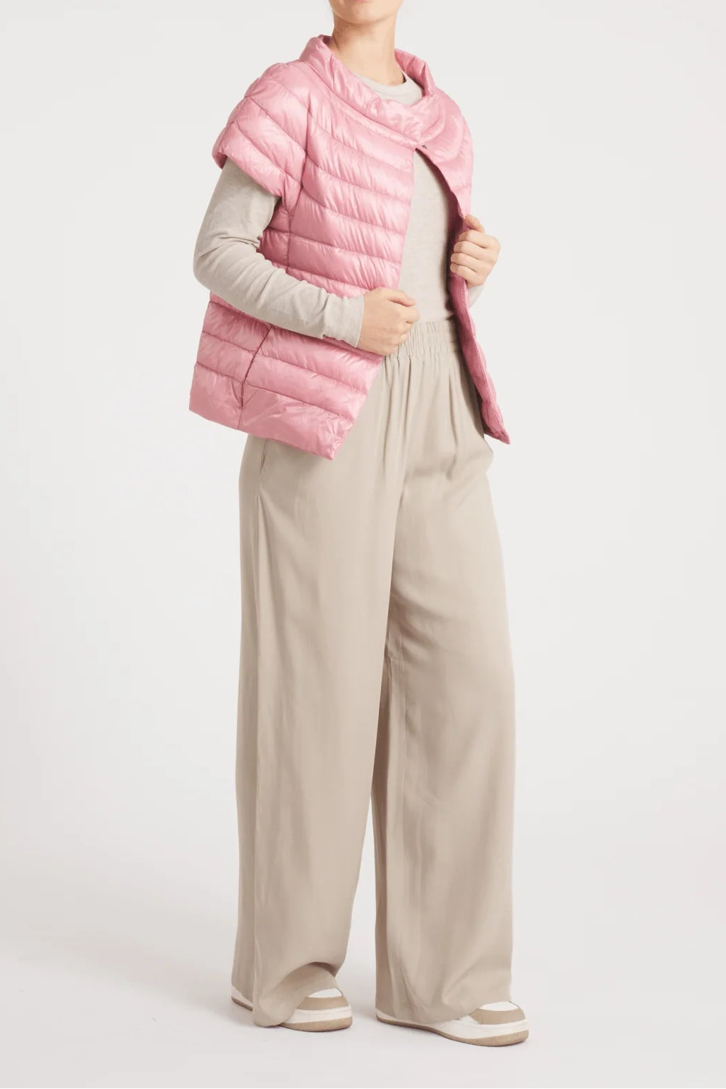Person wearing a pink puffer vest over a light gray long-sleeve shirt and beige pants on a white background