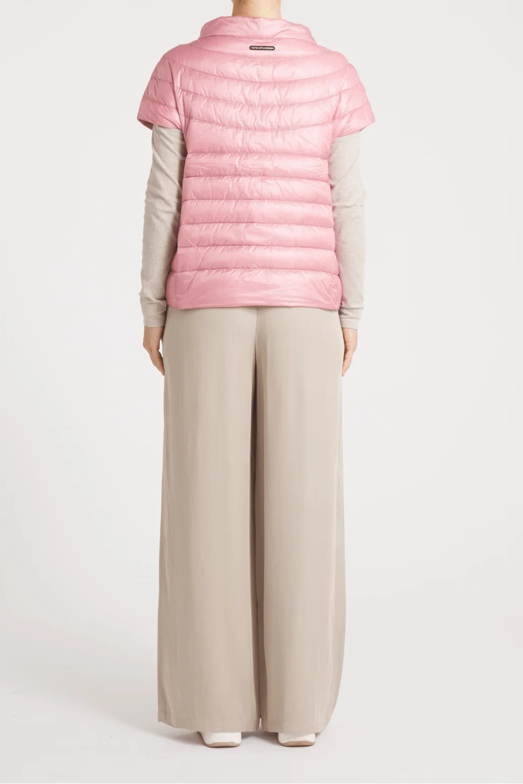 A woman viewed from the back wearing a light pink short-sleeve puffer vest over a beige long-sleeve top and beige wide-leg trousers, against a plain light gray studio background.