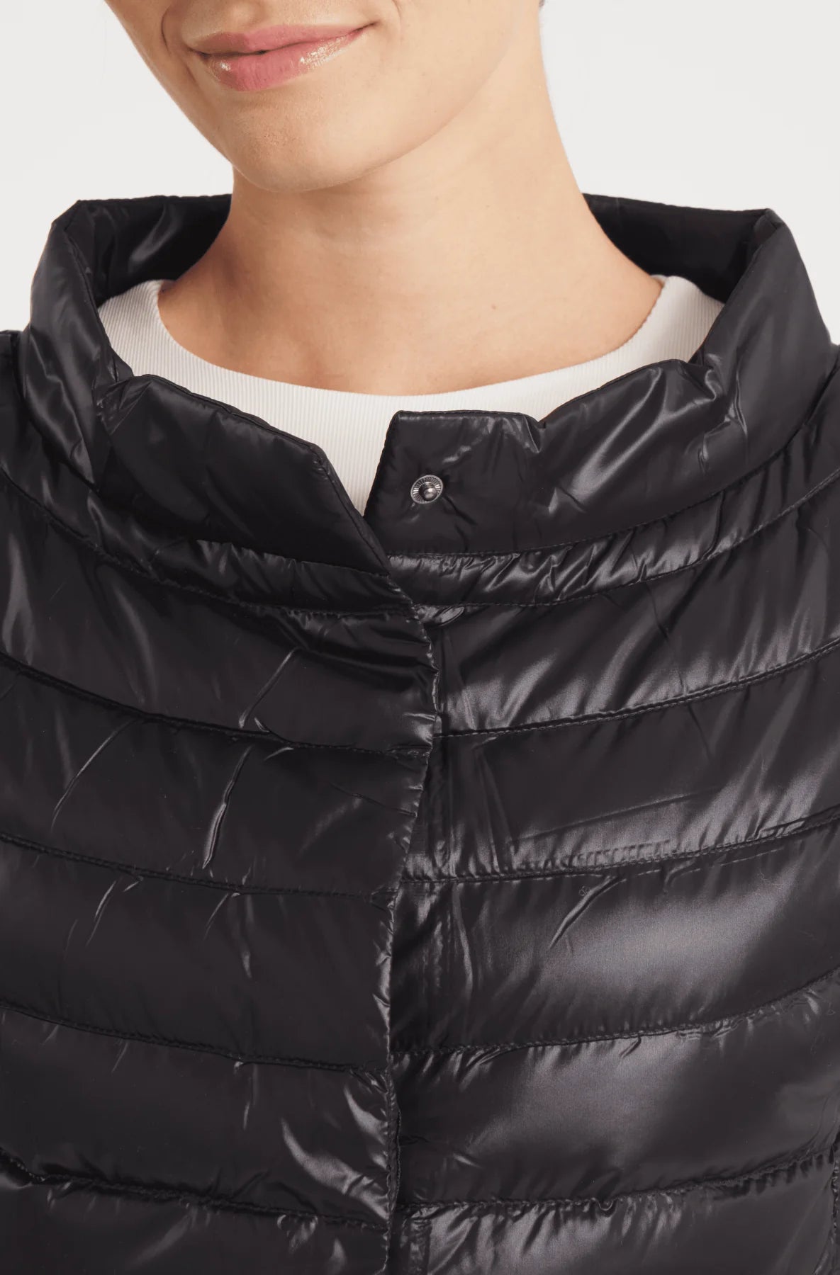 Close-up of a person wearing a shiny black quilted puffer jacket with a high collar over a white shirt, against a light gray background.