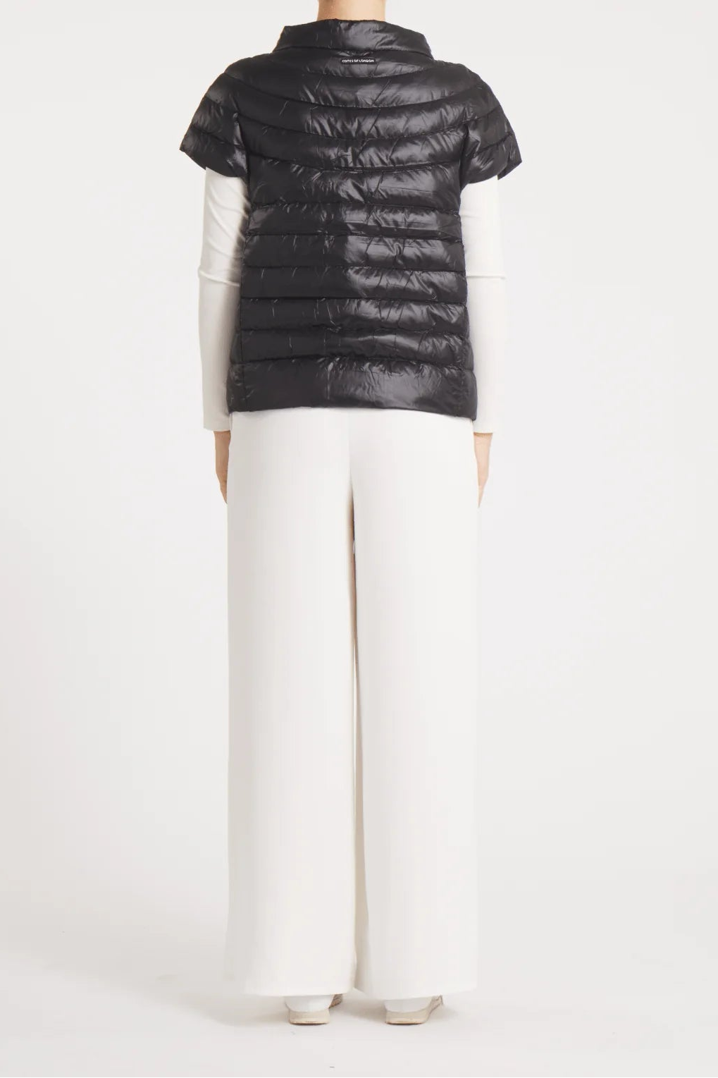A model shown from the back wearing a black short-sleeve puffer vest over a white long-sleeve top and white wide-leg trousers, set against a plain white studio background.