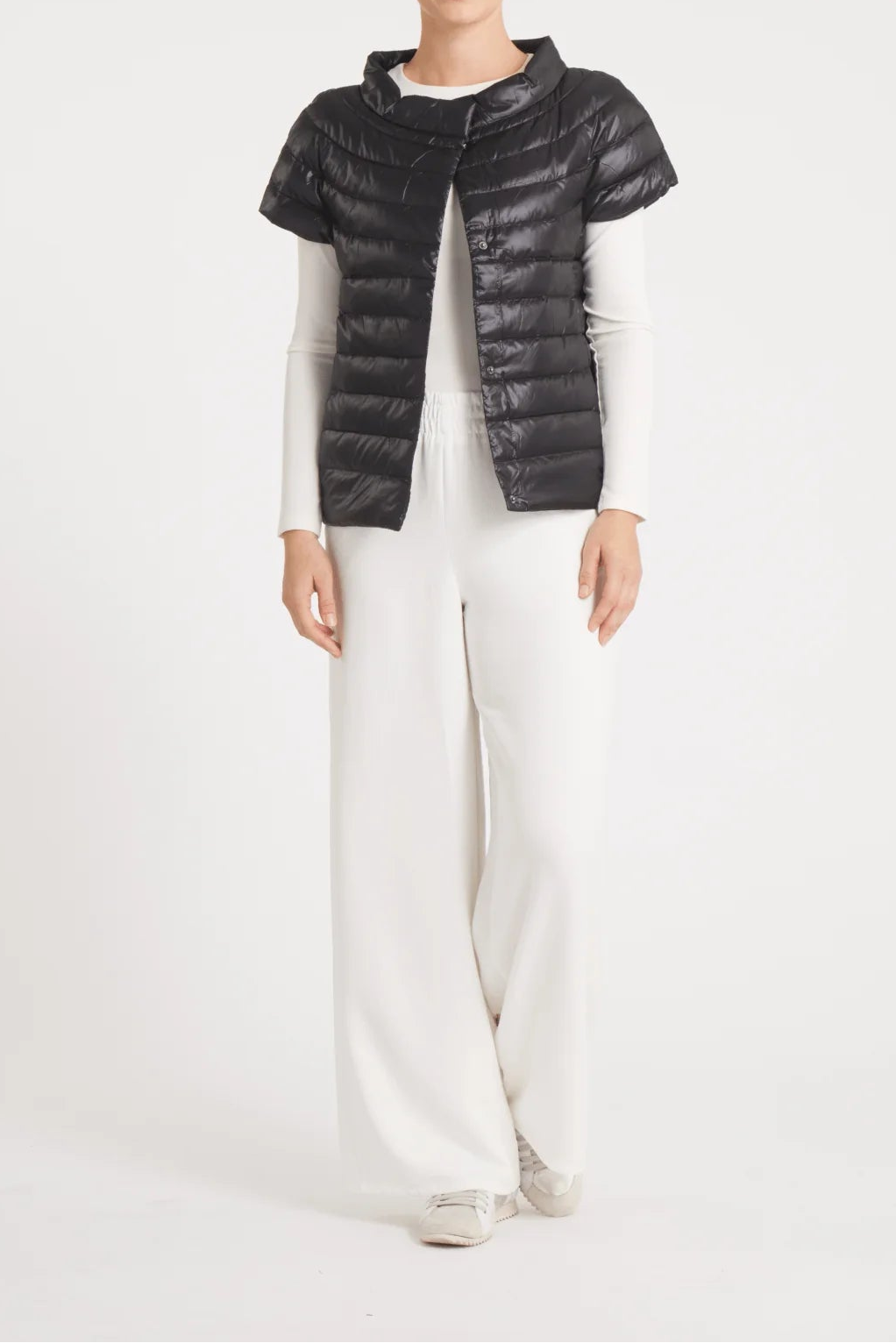 A model stands against a plain white studio background wearing a black short-sleeve puffer vest over a white long-sleeve top and white wide-leg trousers.