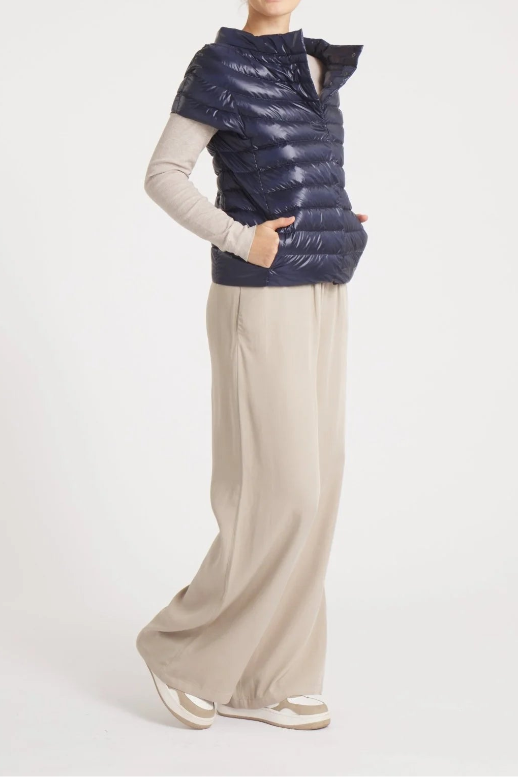 Person wearing a blue puffer vest over a light gray long-sleeve shirt and beige pants on a white background