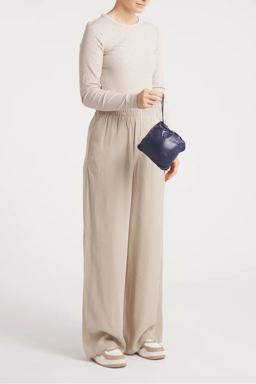 A woman modeling a light beige long-sleeve top and matching wide-leg trousers, holding a small dark blue handbag against a plain light gray studio background.