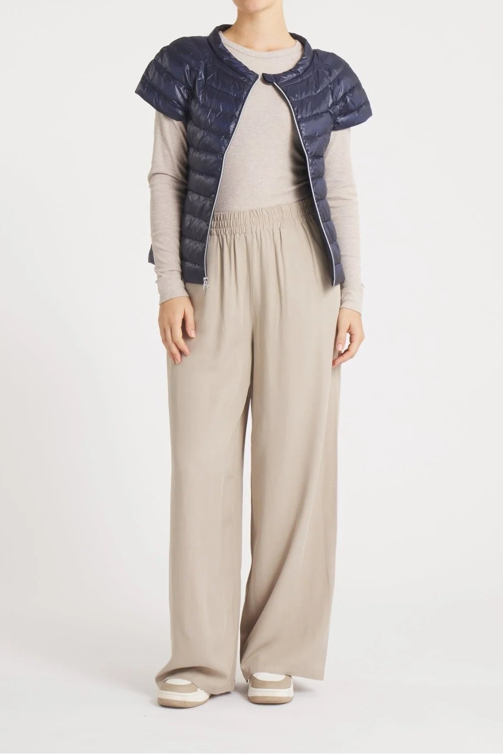 A model stands against a light gray studio background wearing a navy blue quilted puffer vest with short sleeves over a beige long-sleeve top and matching beige wide-leg trousers.