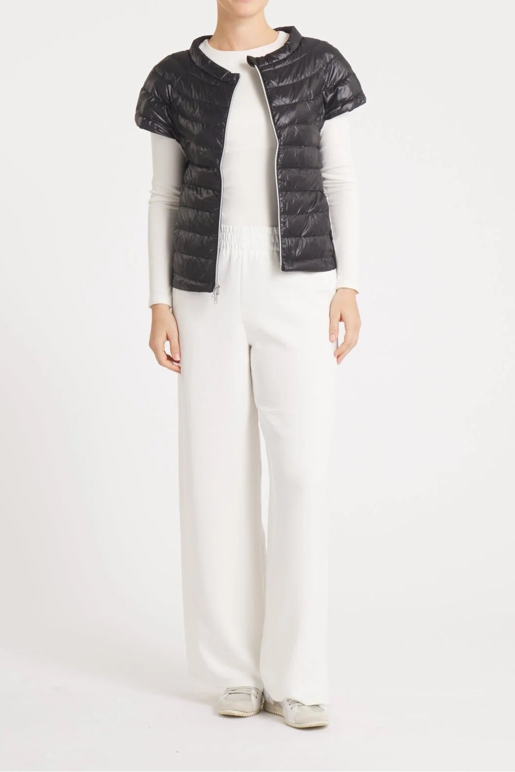 A model stands against a plain white studio background wearing a black quilted puffer vest over a white long-sleeve top and white wide-leg trousers.