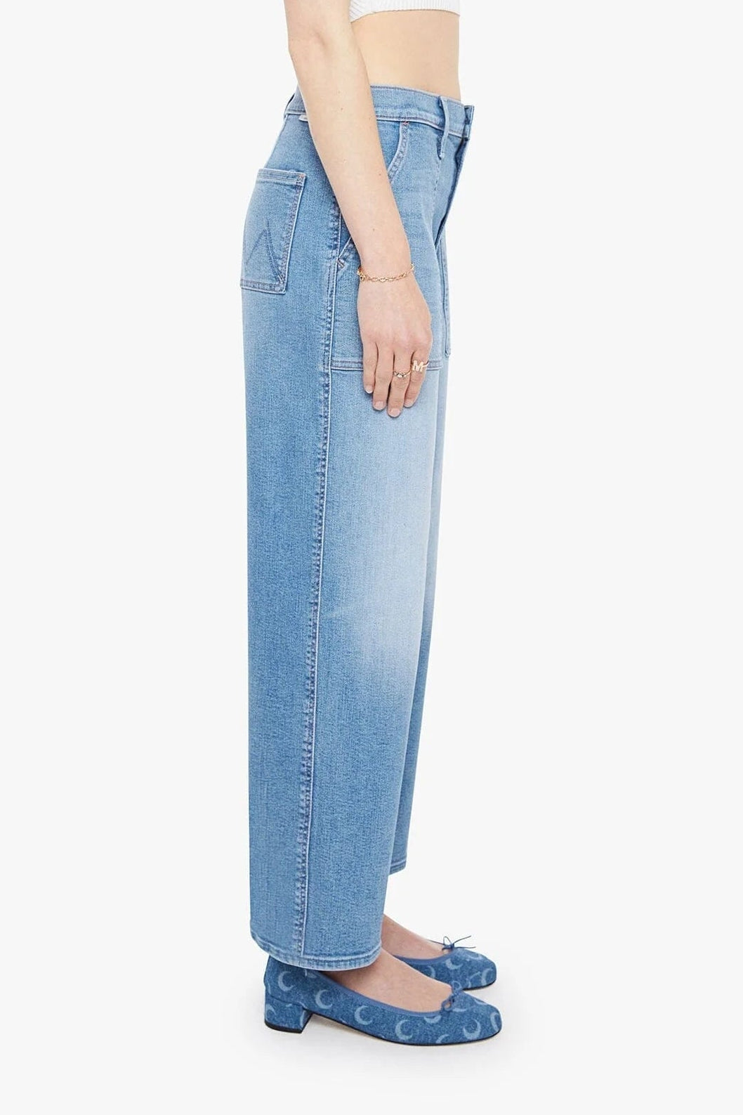 A side-profile view of a model wearing light blue wide-leg denim jeans, a white crop top, and blue patterned flats against a plain white background.