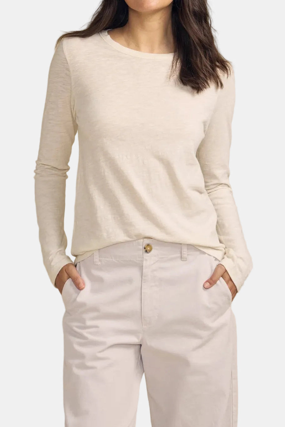 A woman modeling a cream-colored long-sleeve knit top with white pants, standing against a solid beige studio background.