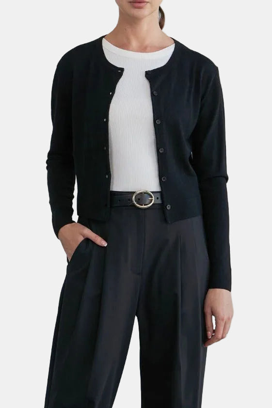 A woman stands in a studio wearing a black cardigan over a white top with black wide-leg trousers and pointed shoes.