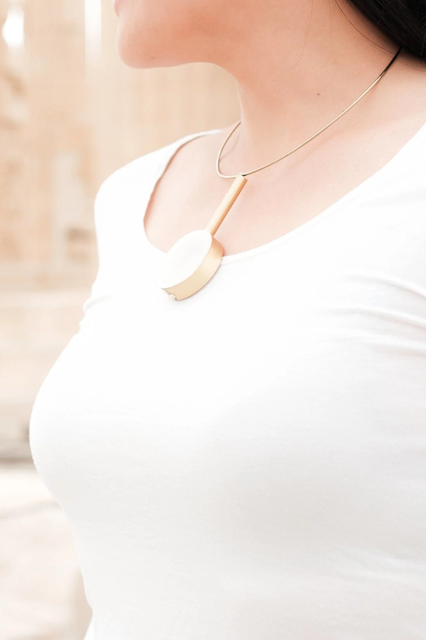 A close-up lifestyle shot of a woman wearing a modern gold pendant necklace with a large, flat circular disc and a thin vertical bar, paired with a white top.